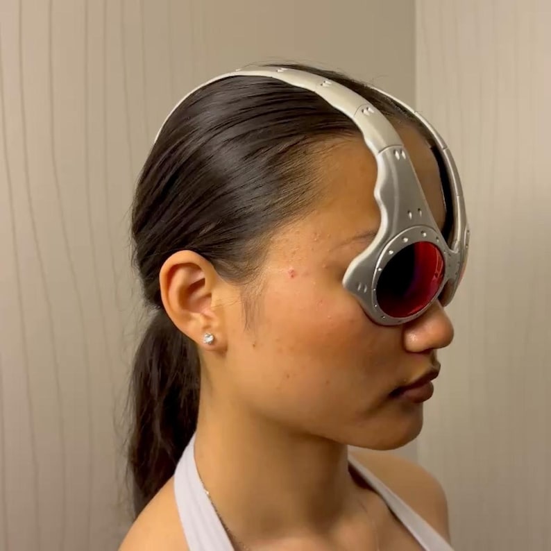 May include: Silver and red goggles with a curved, metallic frame and round lenses. The goggles have a silver headband and are worn on the head. The lenses are a deep red color.