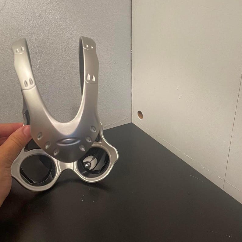 May include: Silver-toned sunglasses with a unique, futuristic design. The sunglasses feature a wide, curved top piece and circular lenses. The frame has a matte finish, and the lenses appear dark. The overall style is modern and sporty.