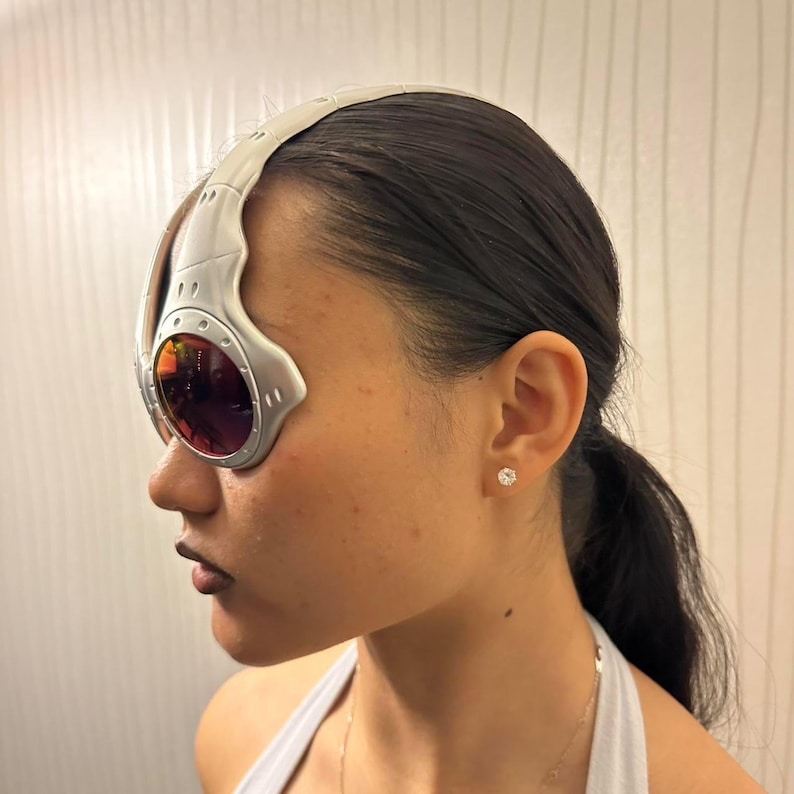 May include: Silver-framed sunglasses with red-tinted lenses. The sunglasses have a unique, futuristic design with a curved top piece and circular lens frames. The lenses are reflective, and the frame is a matte silver color.