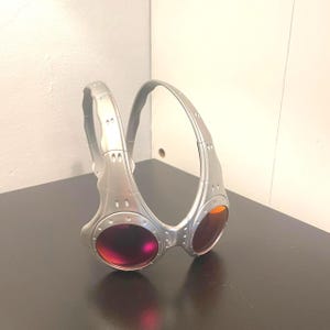 May include: Silver-framed goggles with red and orange lenses. The goggles have a curved, wraparound design with a thick frame and a bridge connecting the two lenses. The lenses are oval-shaped and the frame has a metallic finish.