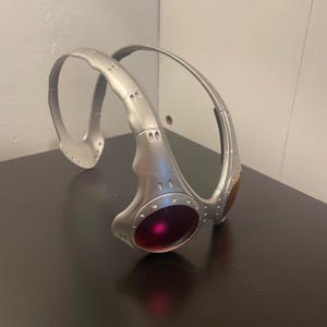 May include: Silver-colored wraparound sunglasses with a futuristic design. The lenses are a deep red and amber color. The frame has a metallic finish with visible screws and rivets, giving it a rugged, industrial aesthetic.