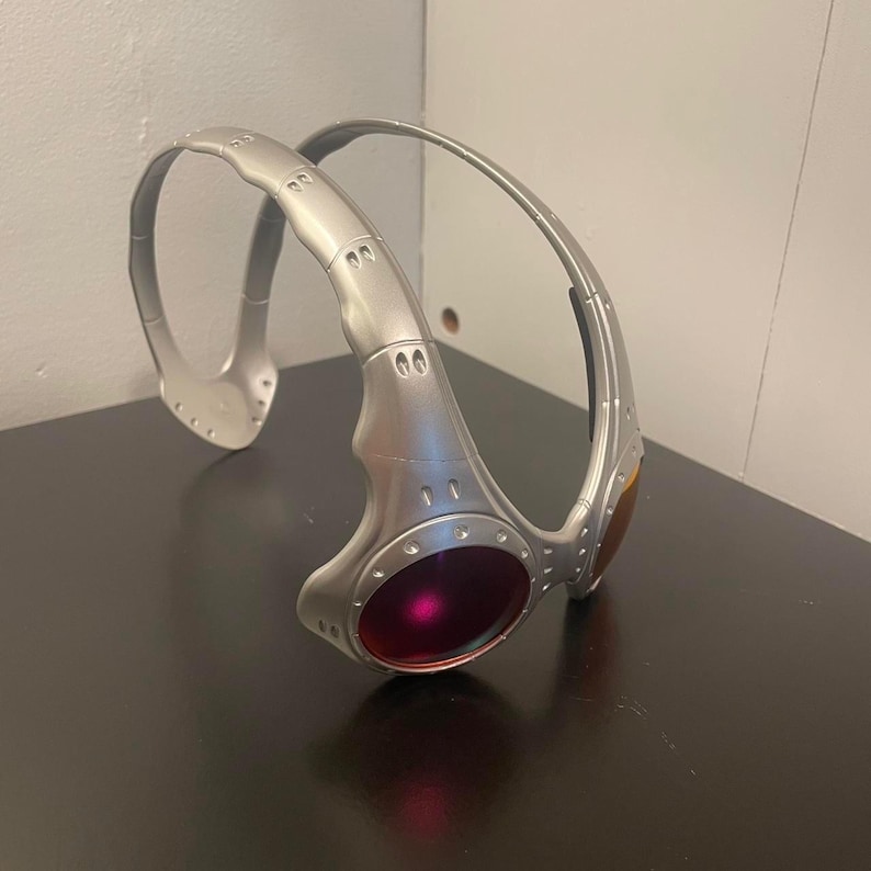 May include: Silver-colored wraparound sunglasses with a futuristic design. The lenses are a deep red and amber color. The frame has a metallic finish with visible screws and rivets, giving it a rugged, industrial aesthetic.