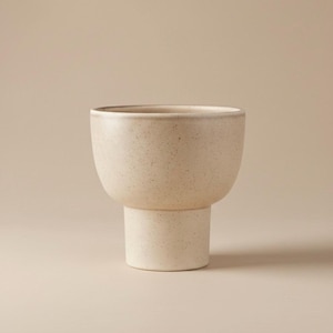 Puede incluir: Una maceta de cerámica moteada de color blanco roto, con una parte superior redondeada y una base cilíndrica. La maceta tiene un diseño moderno y minimalista, adecuado para exhibir pequeñas plantas o suculentas. El color neutro complementa varios estilos de interior.
