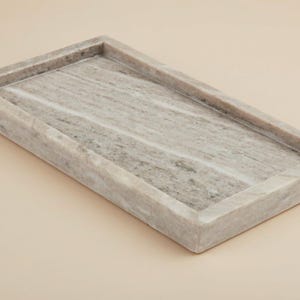 May include: Rectangular marble tray with a raised border. The tray is a light gray color with darker gray veining throughout. The tray is approximately 12 inches long and 6 inches wide.