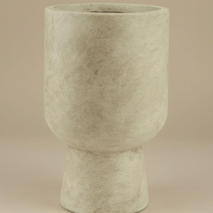 Puede incluir: Una maceta cilíndrica alta de color beige con una parte superior redondeada y una base de pedestal. La superficie texturizada le da un aspecto natural. Adecuada para uso en interiores o exteriores, el diseño es moderno y minimalista. Aproximadamente 60 cm de altura.
