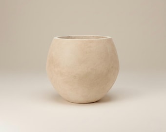 Pode incluir: Um vaso bege em forma de ovo com uma abertura larga. O vaso tem uma superfície texturizada e uma base arredondada. A cor neutra e o design simples tornam-no adequado para várias plantas e estilos de decoração.