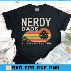 May include: Black t-shirt with the text "NERDY DADS BUILD CHARACTER" in a distressed font. The design includes a retro-style graphic with a multi-colored sunburst and a geometric shape. White sneakers and blue jeans are also visible.