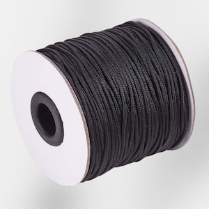 May include: A spool of black nylon cord. The cord is tightly wound around a white cardboard core. The black cord has a smooth, slightly shiny appearance. The spool is set against a plain, light background.