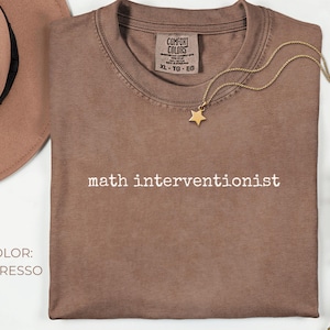 May include: Espresso-colored t-shirt with the text "math interventionist" in white. A gold star pendant necklace rests on the shirt. A brown hat and sprigs of pink flowers are in the background.