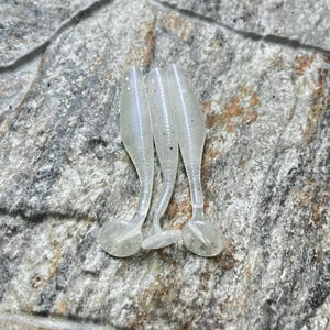 May include: Three clear, soft plastic fishing lures with a pearlescent shimmer. Each lure has a curved body, a small tail, and a rounded head. The lures are arranged vertically on a textured, gray stone surface.