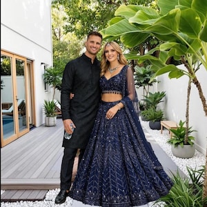 Puede incluir: Una pareja posa al aire libre. La mujer lleva un conjunto formal de dos piezas azul marino adornado con un chal transparente a juego. El hombre viste un kurta negro bordado y pantalones a juego. Están sobre una terraza de madera.