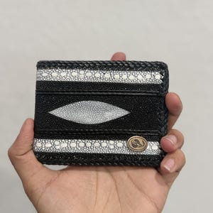 May include: A black and white stingray leather wallet with an oval design. The wallet has white dotted stripes and a metal emblem. The edges are braided for detail.