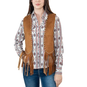May include: A woman wearing a brown suede vest with fringe and silver studs over a patterned button-down shirt and blue jeans. The shirt has a white background with red, black, and blue geometric patterns. The vest is a medium brown color.
