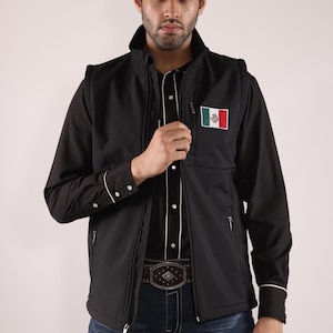 May include: Black vest with a Mexican flag patch, worn over a black shirt with white trim and a decorative belt. The vest has a zipper closure and a stand-up collar. The person is wearing blue jeans.