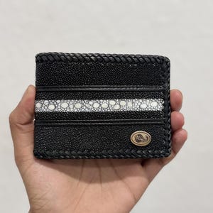 Peut inclure: Portefeuille en cuir noir avec un motif rayé en raie blanche et noire. Le portefeuille a une bordure tressée et un petit emblème ovale. Le portefeuille est tenu dans une main.