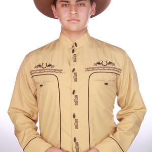 Puede incluir: Camisa de vaquero color beige con ribetes y bordados marrón oscuro. La camisa tiene cuello mandarín y cierre de botones. Un sombrero de vaquero marrón y un cinturón decorativo completan el conjunto.