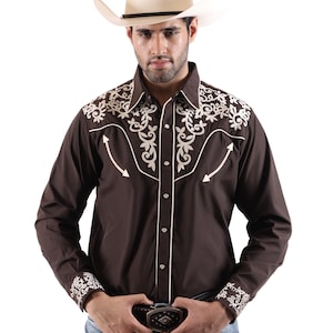 May include: A brown Western-style shirt with white embroidered floral designs on the shoulders and cuffs. The shirt has a button-down front and is paired with a light-colored cowboy hat and blue jeans. A black belt with a decorative buckle is also visible.