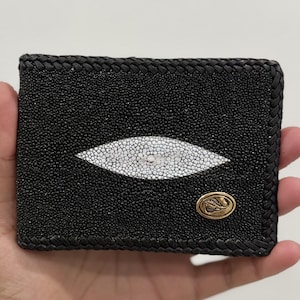 Könnte beinhalten: Schwarzes Lederportemonnaie mit einem weißen, augenförmigen Design in der Mitte. Das Portemonnaie hat einen geflochtenen Rand und ein goldfarbenes Emblem. Das Portemonnaie wird in einer Hand gehalten.