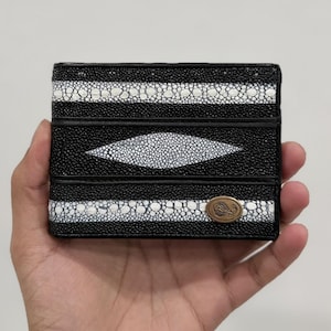 May include: A black and white leather wallet with a distinctive pattern. The wallet has a central diamond-shaped design and horizontal stripes. A small, oval emblem is on the bottom right. The wallet is held in a hand.