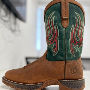 Puede incluir: Bota de vaquero de cuero marrón con caña verde con costuras decorativas rojas y blancas. La bota tiene suela y tacón marrones, y la parte superior de la caña está ribeteada en cuero marrón. El texto "THE AMERICAN WEST" es visible.