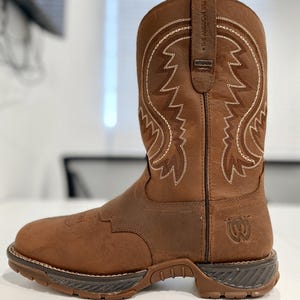 以下が含まれることがあります： 白いステッチと頑丈なソールを備えた茶色の革のカウボーイブーツ。 ブーツには装飾的なステッチパターンがあり、プルストラップには「THE AMERICAN WEST」の文字があります。 ブーツには防水ラベルが付いています。