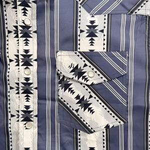 May include: A blue button-up shirt with a vertical striped pattern. The shirt features a white and black geometric design on the front, pocket, and cuffs. The shirt has silver buttons and a classic collar.