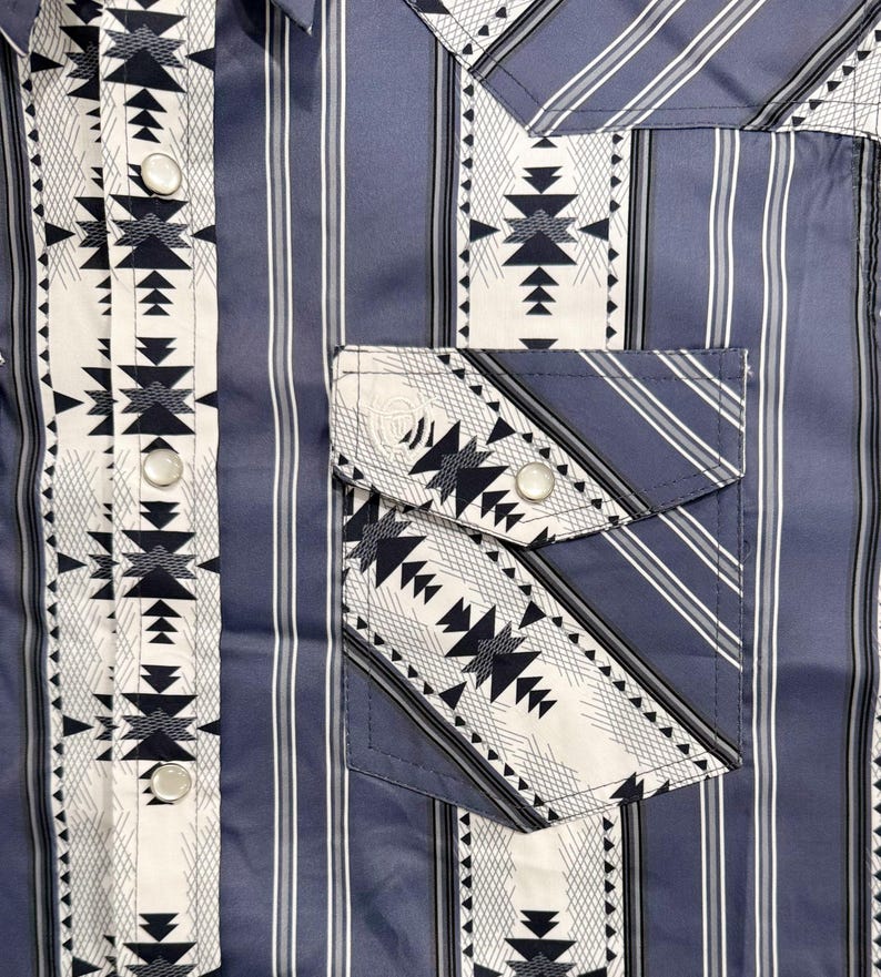 May include: A blue button-up shirt with a vertical striped pattern. The shirt features a white and black geometric design on the front, pocket, and cuffs. The shirt has silver buttons and a classic collar.