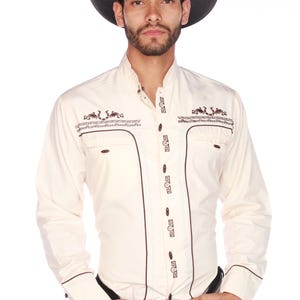 Puede incluir: Camisa vaquera color crema con detalles y ribetes bordados en marrón. La camisa tiene cuello mandarín, cierre de botones y dos bolsillos en el pecho. Un sombrero de vaquero negro completa el look.