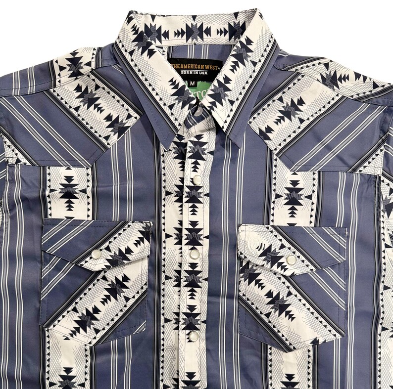 May include: A blue and white patterned button-up shirt with a Western design. The shirt features vertical stripes and geometric patterns on the pockets, collar, and yoke. The text "THE AMERICAN WEST - BORN IN USA" is visible on the collar.