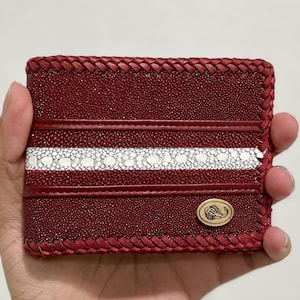 May include: A burgundy leather wallet with a braided edge and a white and burgundy stripe design. The wallet has a small, oval-shaped emblem with a stylized design. The wallet is held in a hand.