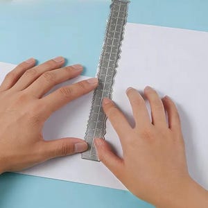 May include: A silver metal ruler with decorative edges is held over a white piece of paper on a light blue surface. The ruler has measurements marked along its length. The hands are holding the ruler and paper.
