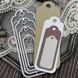 Könnte beinhalten: Eine Sammlung von Metall-Stanzformen in verschiedenen Größen, in Form von Anhängern mit dekorativen Kanten. Mehrere geschichtete Papieranhänger in Braun-, Weiß- und Mauve-Tönen sind ebenfalls vorhanden, passend für Bastel- oder Scrapbooking-Projekte. Die Anhänger haben ein Loch oben.