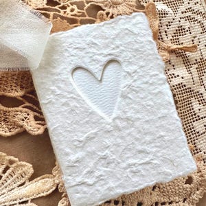 May include: A rectangular white handmade paper card featuring a die-cut heart shape in the centre. The paper has a thick, textured, deckled edge finish. A sheer white ribbon is attached to the top left corner of the card.