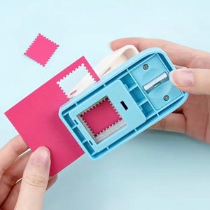 Puede incluir: Un troquelador de papel azul claro con forma de sello y un mango blanco. Se está perforando un trozo de papel rosa, creando un recorte con forma de sello y un sello rosa. El fondo es azul claro.