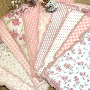 Könnte beinhalten: Eine Sammlung von rosa und weiß gemusterten Papierbögen mit gezackten Kanten. Die Designs umfassen Blumen-, Tupfen-, Streifen- und Karomuster. Getrocknete weiße Blumen sind auf den Papierbögen platziert.
