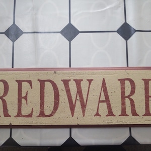 May include: A rectangular wooden sign with the word "REDWARE" in red, distressed lettering. The background is a cream color with a crackled effect, and the edges are a darker red. The sign is likely for kitchen or home decor.