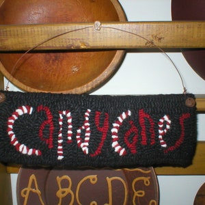 May include: A black and white needlepoint sign with the word "Candy Cane" in red and white stripes. The sign is hanging from a wooden shelf with two wooden bowls on either side.