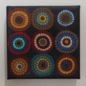 May include: A square canvas painting featuring a grid of nine colorful concentric circles. Each circle is segmented with mosaic-like patterns in shades of red, orange, yellow, blue, and green against a black background. The artwork is mounted on a black frame.