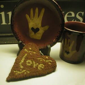 May include: A brown heart-shaped rug with the word "Love" woven into it. The rug is sitting on a table next to a red ceramic mug and a red ceramic plate with a hand painted on it.