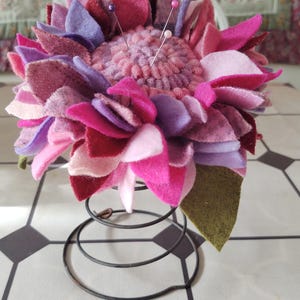 May include: A pink and purple felt flower pincushion with a spring base. The pincushion is made of many layers of felt petals and has several pins sticking out of it.