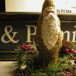 May include: A green felt Santa Claus figure with a white beard and a wreath of pine branches, red berries, and pine cones.