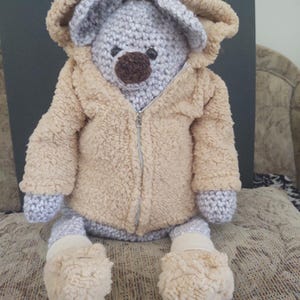 May include: A handmade crochet teddy bear wearing a tan hooded jacket with a zipper. The bear has gray ears, a brown nose, and gray paws and feet. The bear is sitting on a patterned surface.
