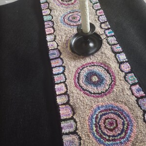 May include: A hand-hooked table runner with a neutral background and colorful circular designs. The runner features a repeating pattern of concentric circles in shades of blue, purple, and pink. A black candle holder with a white candle sits on the runner.