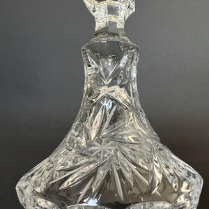 May include: A clear cut glass candlestick holder featuring a wide, flared base with intricate starburst and fan-shaped patterns etched into the surface. The stem transitions into a faceted neck, showcasing reflective light refractions throughout.