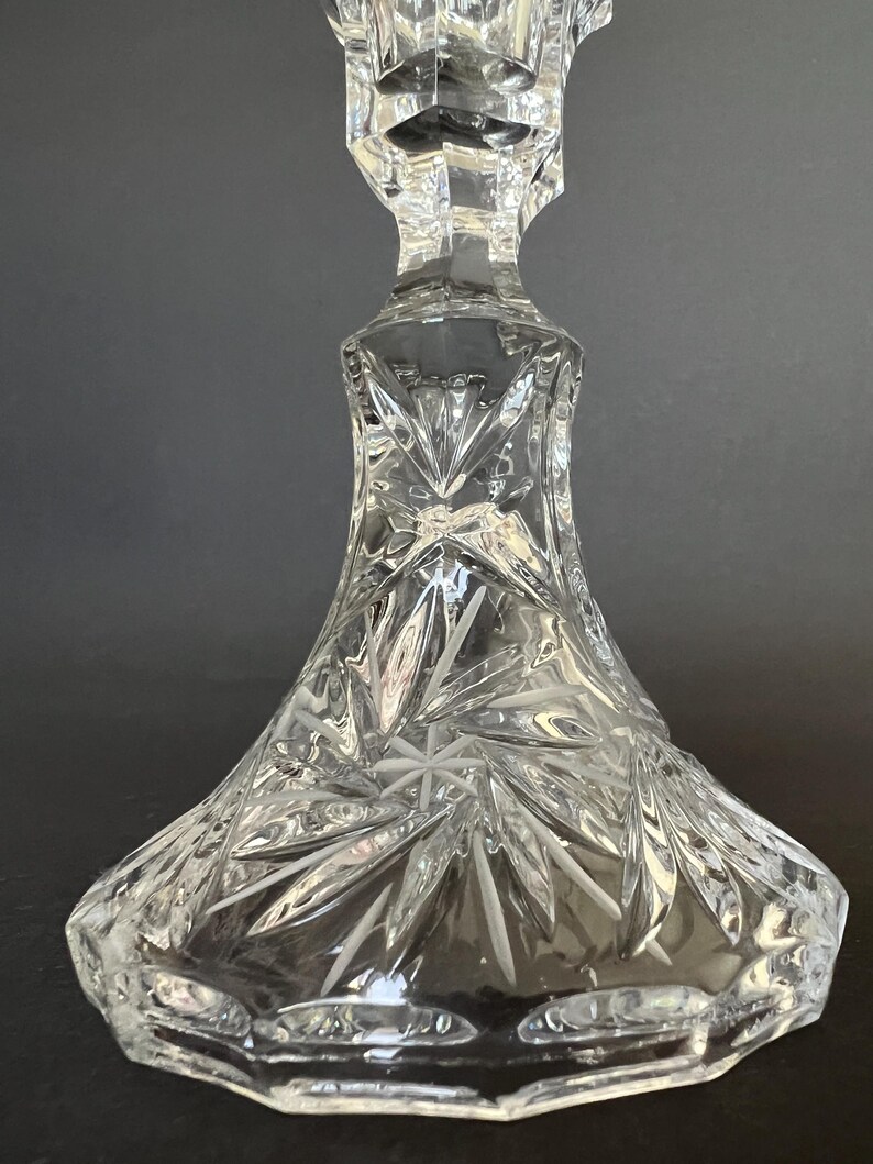 May include: A clear cut glass candlestick holder featuring a wide, flared base with intricate starburst and fan-shaped patterns etched into the surface. The stem transitions into a faceted neck, showcasing reflective light refractions throughout.