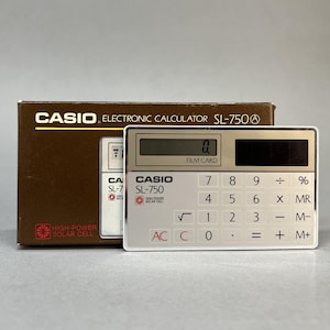 Könnte beinhalten: Ein silberner Casio SL-750 elektronischer Filmkarten-Taschenrechner mit Solarzellenpanel, Digitalanzeige und einem Tastenfeld für Zahlen und Funktionen. Er steht vor der originalen braunen Pappschachtel mit dem Casio-Markennamen.