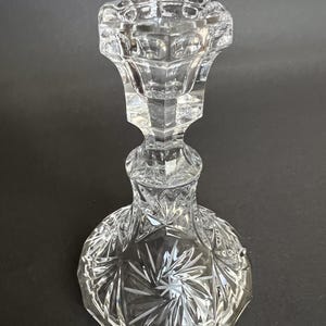 May include: A clear glass candlestick holder featuring a faceted, multi-sided top section and a wide, circular base. The base is decorated with a deeply etched starburst pattern, while the stem and upper cup display geometric, angular cuts.