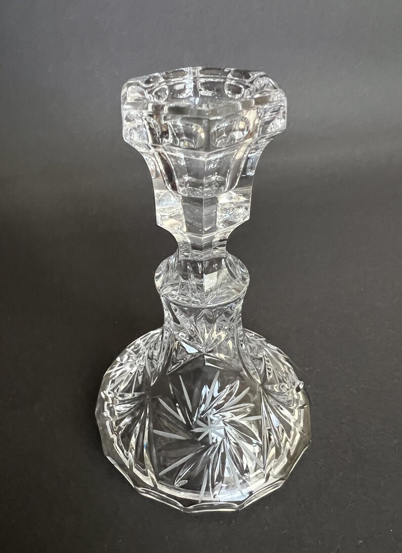 May include: A clear glass candlestick holder featuring a faceted, multi-sided top section and a wide, circular base. The base is decorated with a deeply etched starburst pattern, while the stem and upper cup display geometric, angular cuts.