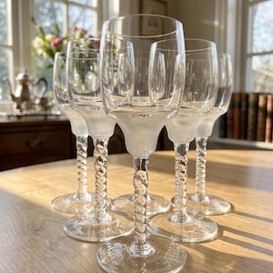 Può includere: Un set di sei calici in vetro trasparente caratterizzati da una coppa liscia e arrotondata, una base satinata e uno stelo decorativo intrecciato. Ogni calice poggia su un piede circolare trasparente, creando un aspetto uniforme per la collezione.