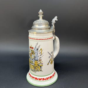 以下が含まれることがあります： 金属製の蓋と親指で開けるレバーが付いた白い陶器製のビールジョッキ。本体にはライオン、旗、交差した剣が描かれたカラフルな紋章があしらわれています。上部と下部の縁には赤と緑の装飾的なボーダーラインが施されています。